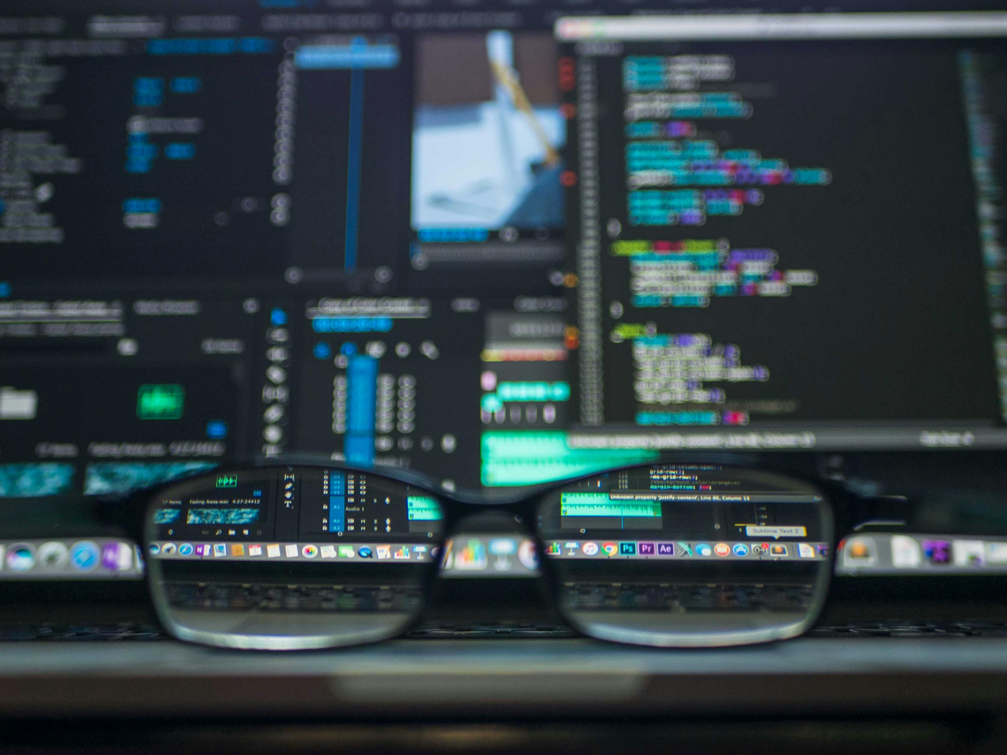 Blurry picture of computer code on a screen with glasses on the desk in front of it